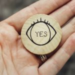 Round wooden coin with eye detail and word, text "yes" printed on it, held in the palm of a woman's hand.
