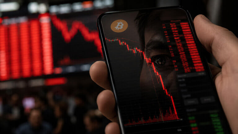 Close-up of a smartphone displaying a falling Bitcoin price chart, overlaid on a red market backdrop with a Bitcoin symbol and blurred cryptocurrency-themed imagery, illustrating a Bitcoin market downturn and price volatility.