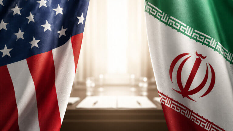 US and Iranian flags facing each other across a negotiation table, symbolizing ongoing talks and uncertainty around a potential US-Iran agreement or ceasefire extension tracked by prediction markets.