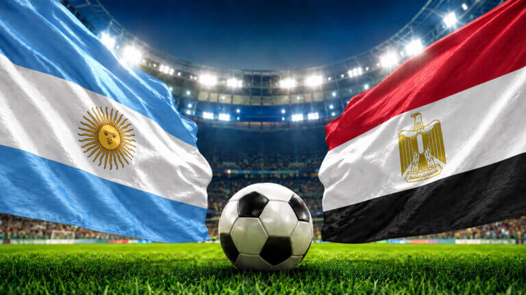 Argentina and Egypt flags facing each other with a soccer ball at midfield inside a brightly lit football stadium