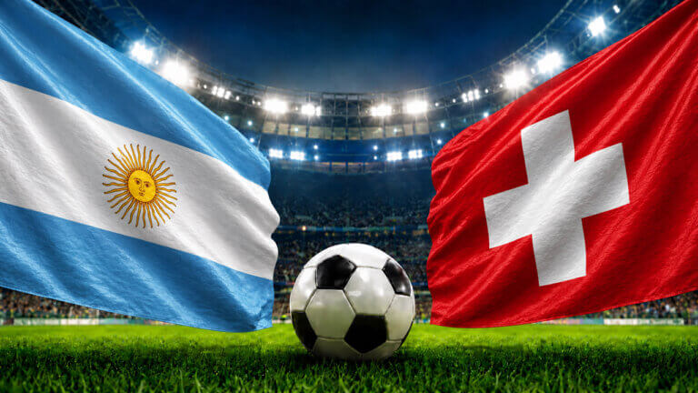Argentina and Switzerland flags wave over a soccer ball on a stadium grass pitch under bright floodlights.