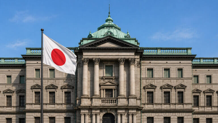Japanese flag waving in front of the Bank of Japan headquarters under a clear blue sky.