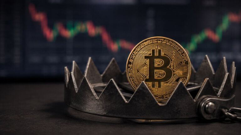 Gold Bitcoin coin caught inside a steel bear trap with a blurred crypto price chart in the background, representing a false bottom signal trapping buyers.