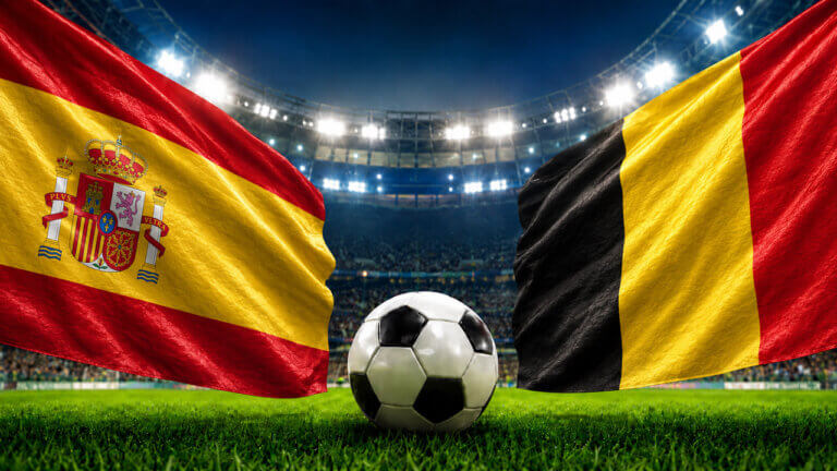 Spain and Belgium flags wave over a soccer ball on a stadium grass pitch under bright floodlights.