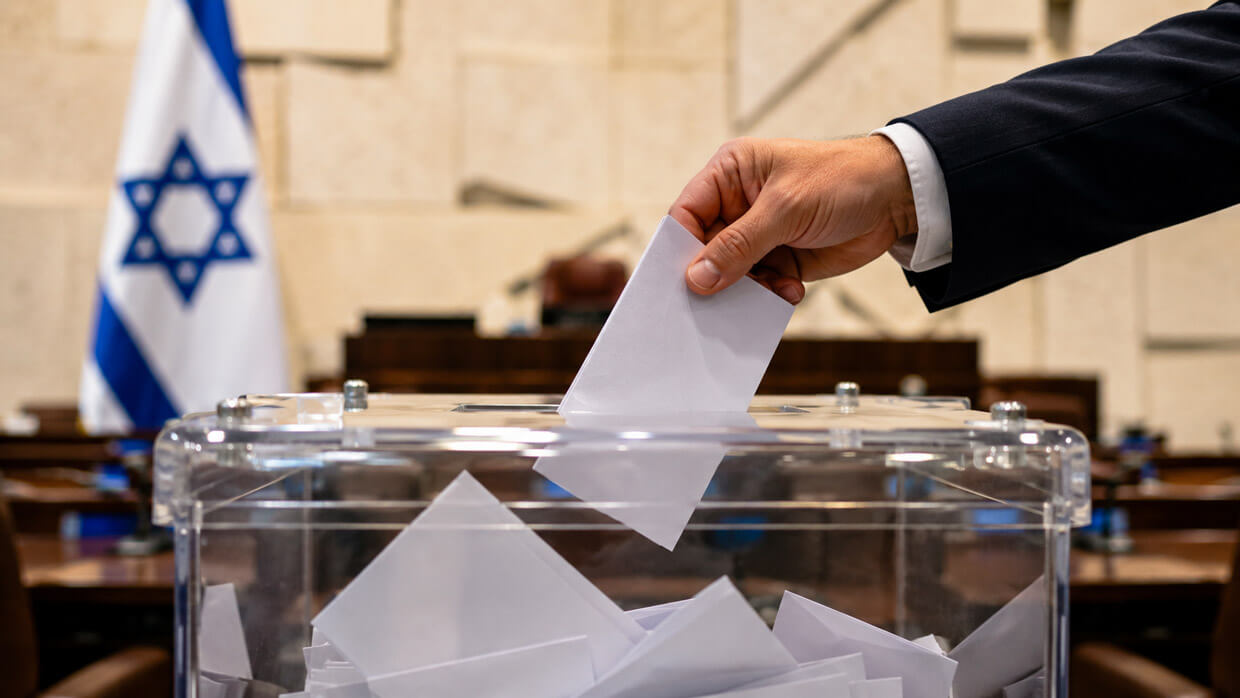Hand placing a blank ballot into a transparent ballot box inside an Israeli government chamber with an Israeli flag in the background.