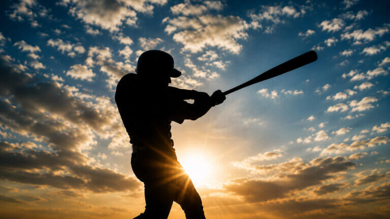 Silhouetted baseball batter completing a powerful swing against a dramatic sunset sky.